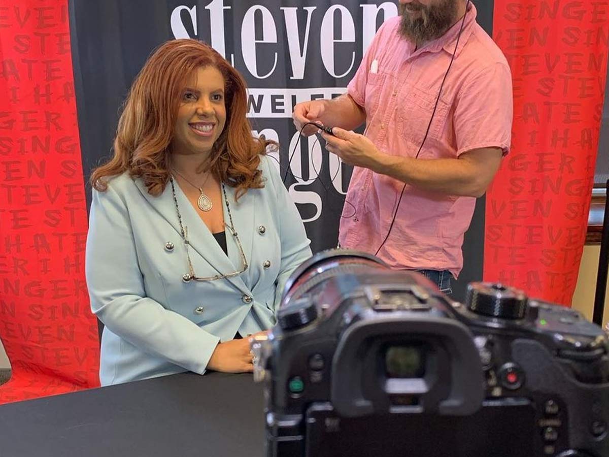 A camera is pointing to a woman in a seat next to a guy with headphones on and a device in his hand.