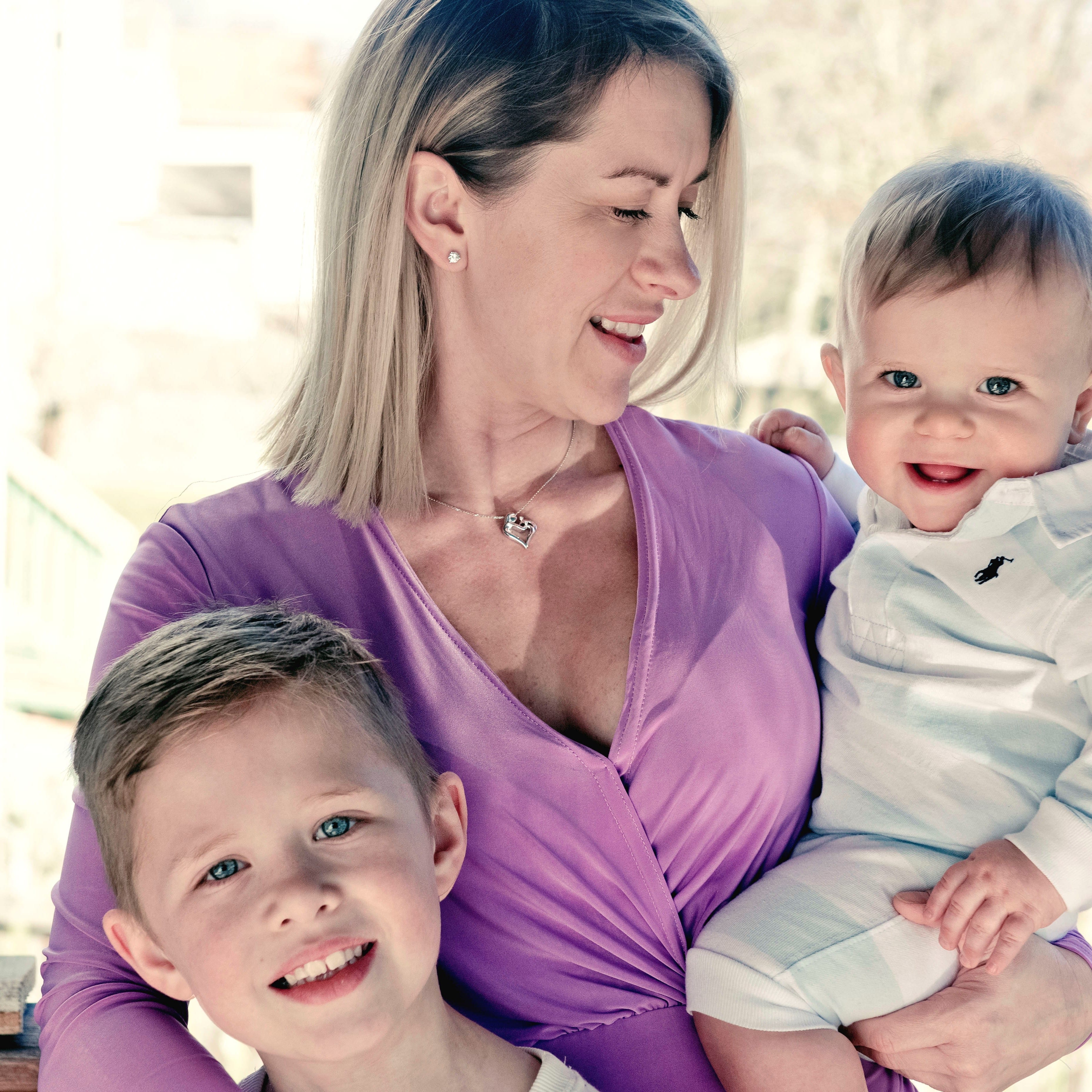 A loving mother holding one of her kids and has her arm around the other with a smile on her face.