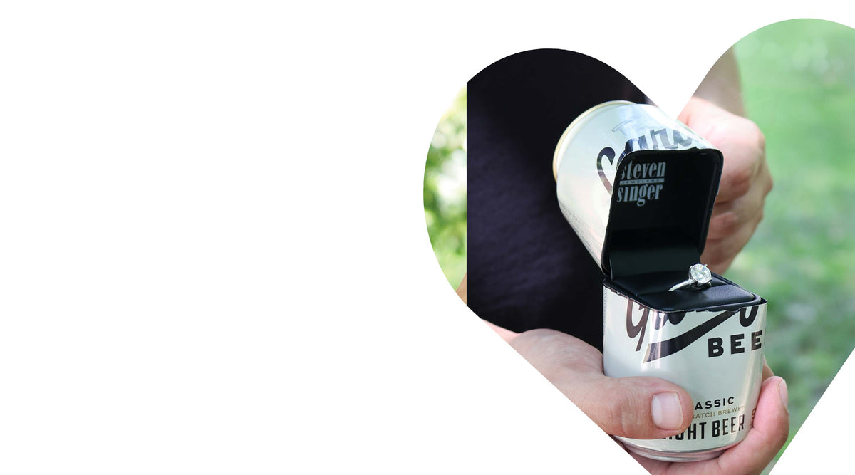 A hand holding an open Steven Singer black ring box inside a can of Garage Beer, with a ring displayed inside, set against a white background with a decorative heart-shaped cutout.