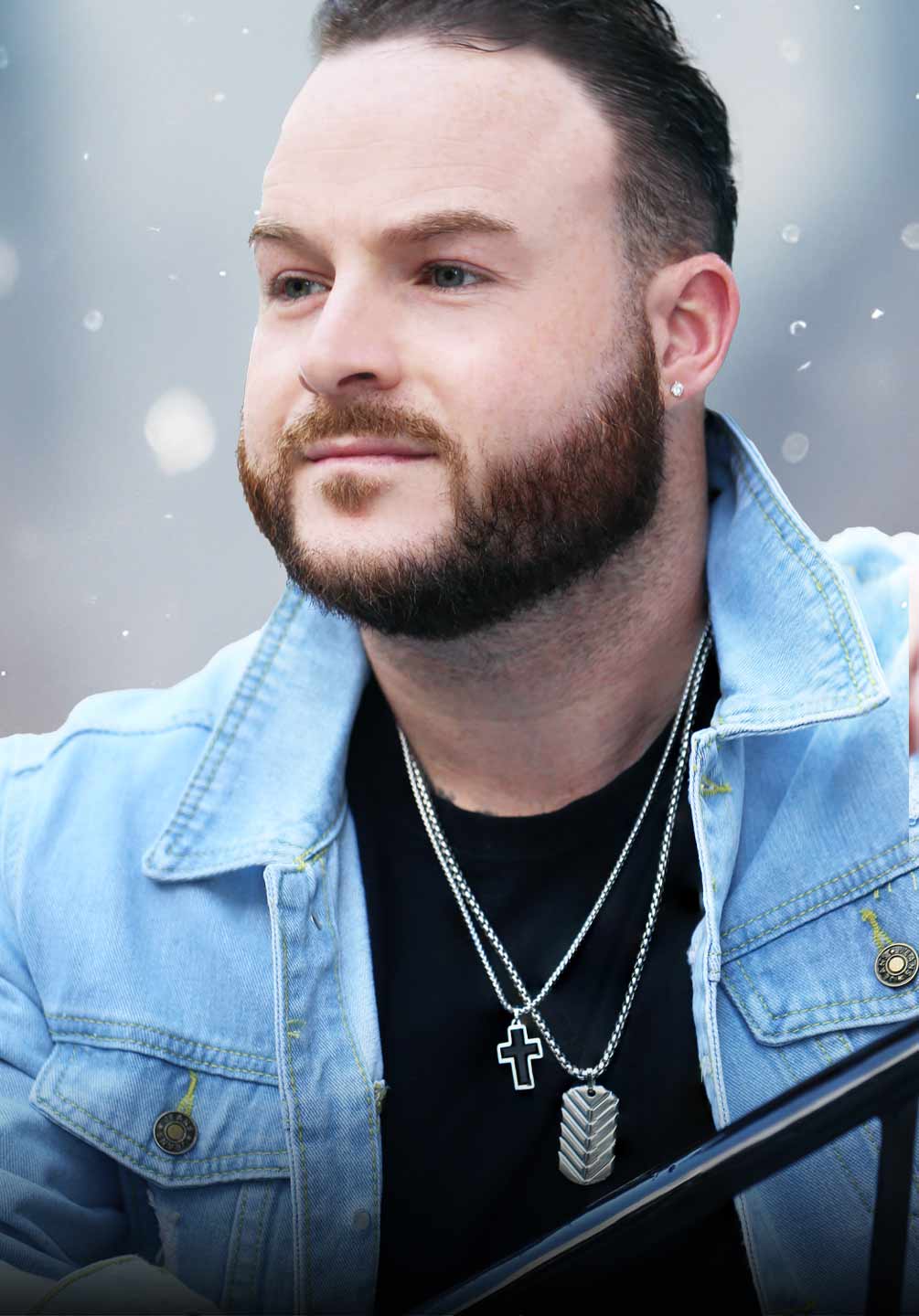A man wearing stylish jewelry including two silver and black necklaces and diamond stud earrings.