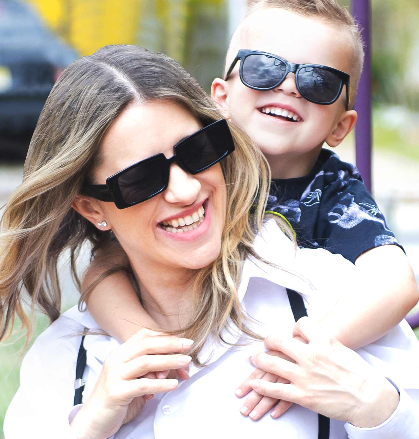 A woman with her son on her back smiling with black sunglasses on.