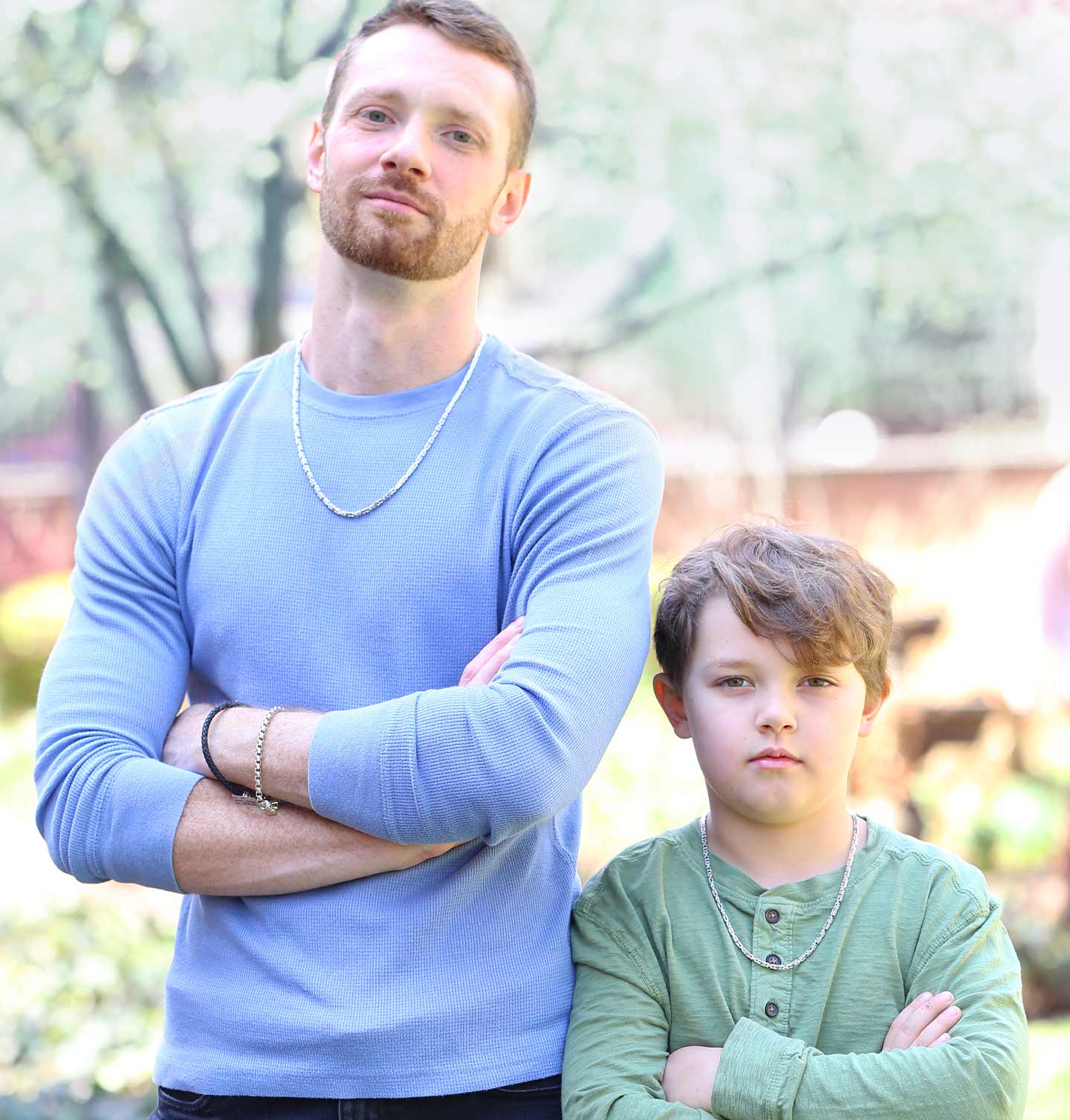 Father and son standing outdoors with arms crossed, wearing light blue and green shirts and chain jewelry.