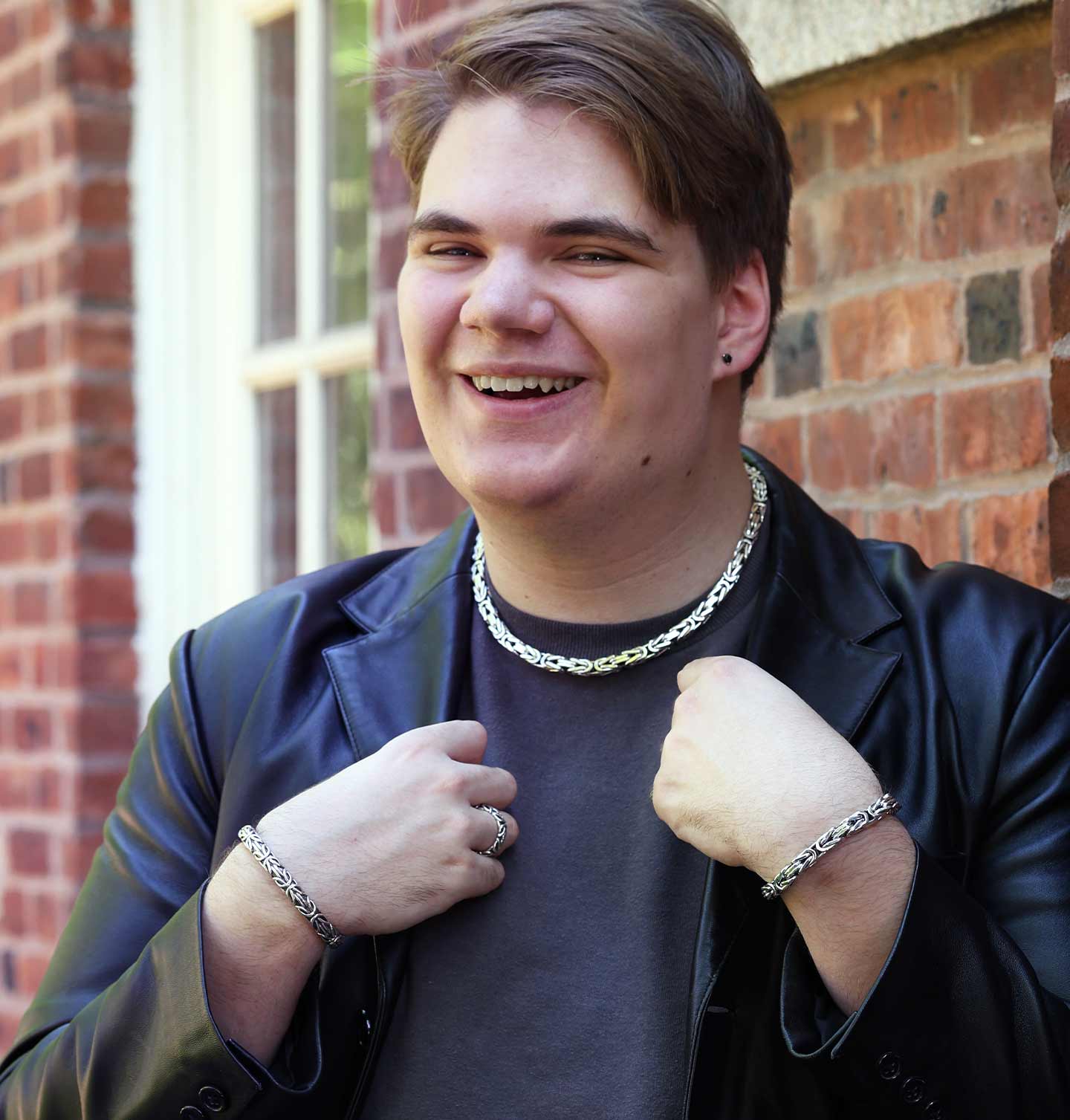Person wearing a black jacket and silver chain necklace in front of a brick wall.