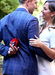 A young man hugging a young woman with his right arm and holding a jewelry box behind his back with his left.