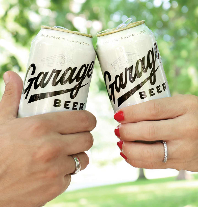 Two hands holding Garage Beer's clinking together with trees in the background.
