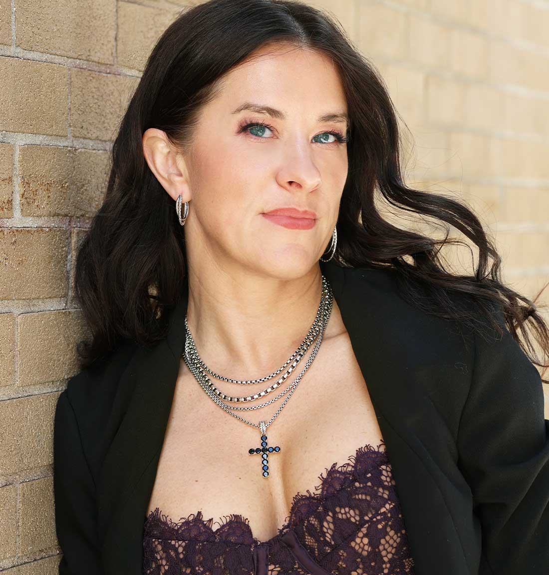 A woman with long dark hair wearing layered silver necklaces, including a cross pendant, a black blazer, and a dark lace top, posing against a beige brick wall.