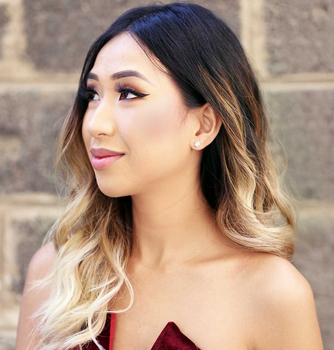 A woman with long, wavy ombre hair transitioning from dark brown to blonde, wearing natural diamond stud earrings and a dark red strapless top, set against a blurred stone wall background.