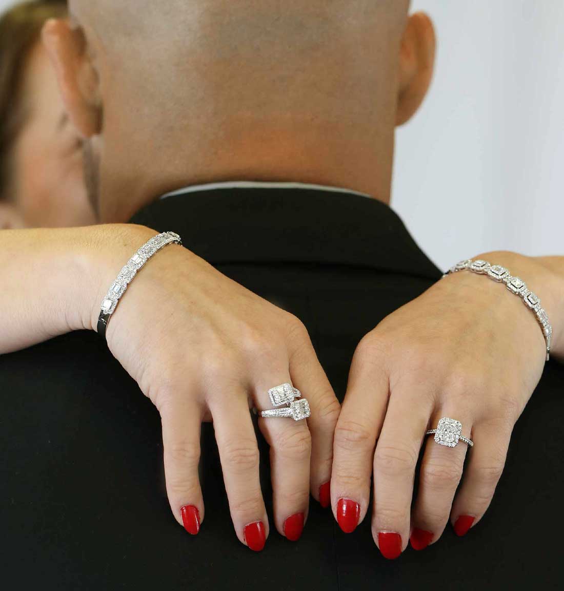 A close-up lifestyle shot of a couple from behind, showing a man in a black suit with a woman's hands resting on his shoulders. The woman is wearing a white gold diamond bracelet on each wrist and engagement rings on both hands, featuring a round-cut center stone and a marquise-cut center stone.