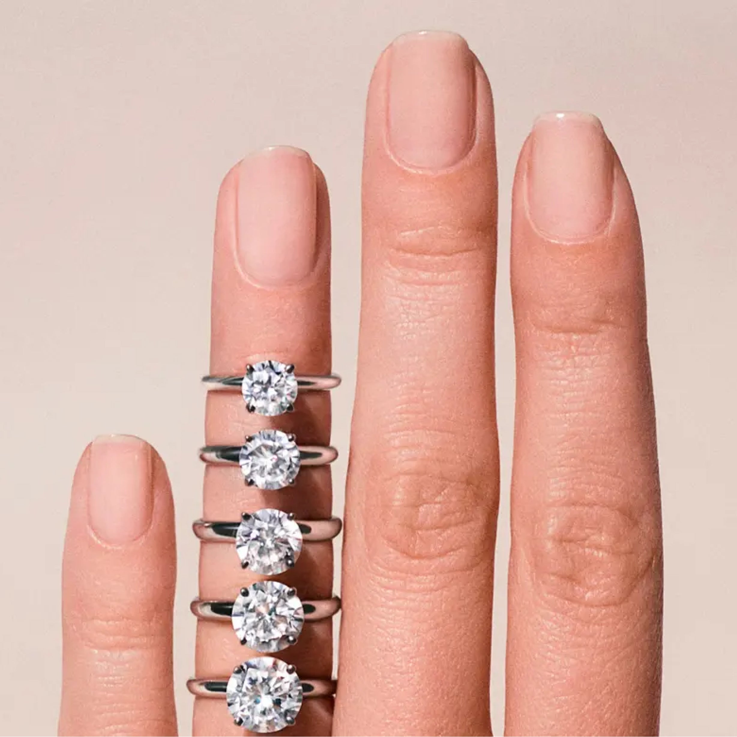 Someone's hand with 5 different sized diamond engagement rings on their ring finger.
